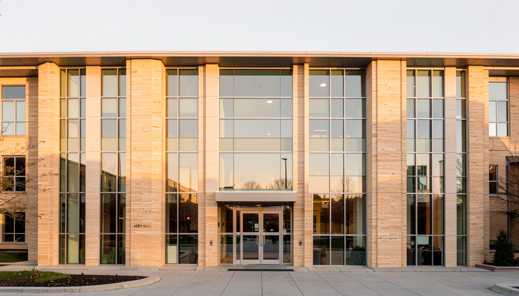 A clean, symmetrical architectural photograph of a modern university library facade with tall glass windows and warm limestone cladding, bathed in soft morning light